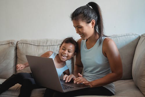 Happy woman with kid sitting on sofa and using laptop while wearing casual clothes in living room