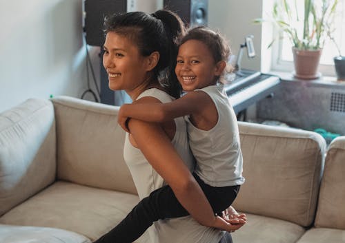 Free Smiling ethnic mother carrying daughter on back while standing near couch in daylight in living room Stock Photo