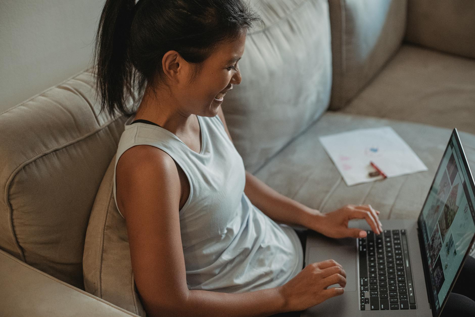 Smiling woman comfortably using laptop on sofa at home, casual and relaxed atmosphere.
