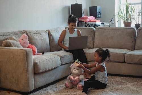 Mother working on computer while sitting on couch and daughter playing with toys on floor in living room