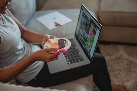 Crop unrecognizable female freelancer using computer in living room while holding plate of plastic food