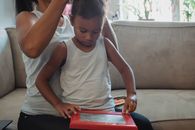 Crop woman in casual outfit sitting on couch and braiding focused little daughter playing with toy