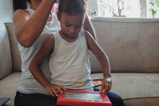 Crop woman in casual outfit sitting on couch and braiding focused little daughter playing with toy