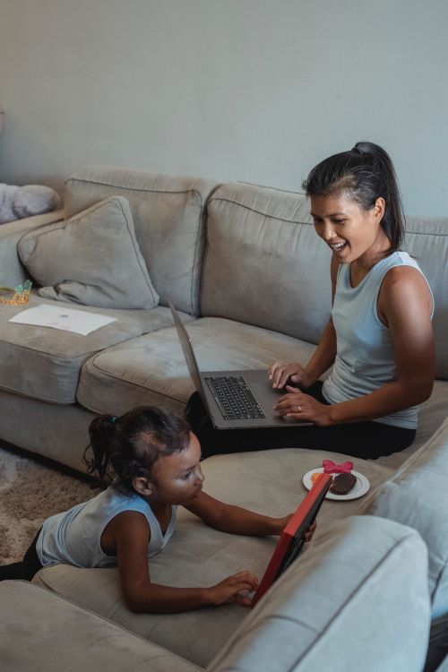 Free Cheerful young Hispanic female looking at cute little daughter playing with toy while working on laptop Stock Photo
