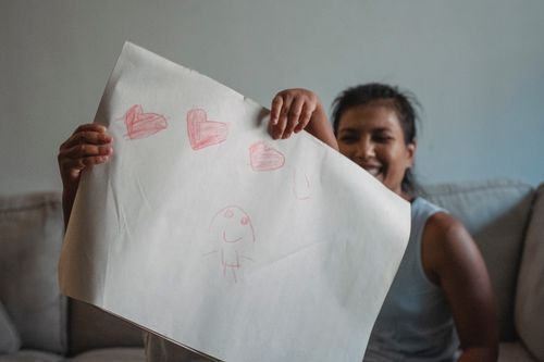 Free Laughing Hispanic female in casual clothes sitting on sofa while daughter showing  paper with drawing Stock Photo
