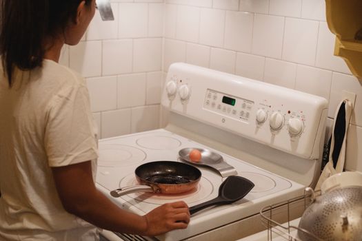 Crop anonymous female in white t shirt standing in kitchen near stove and preparing to cook scrambled eggs