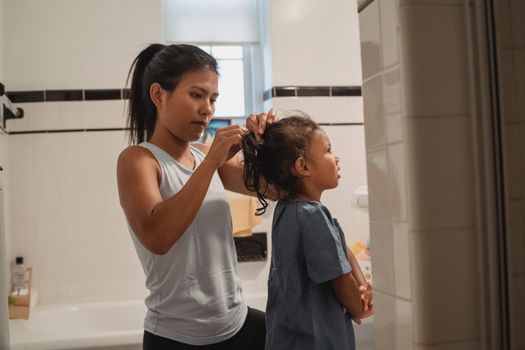 Side view of young Hispanic female in casual outfit standing in bathroom and braiding little daughter