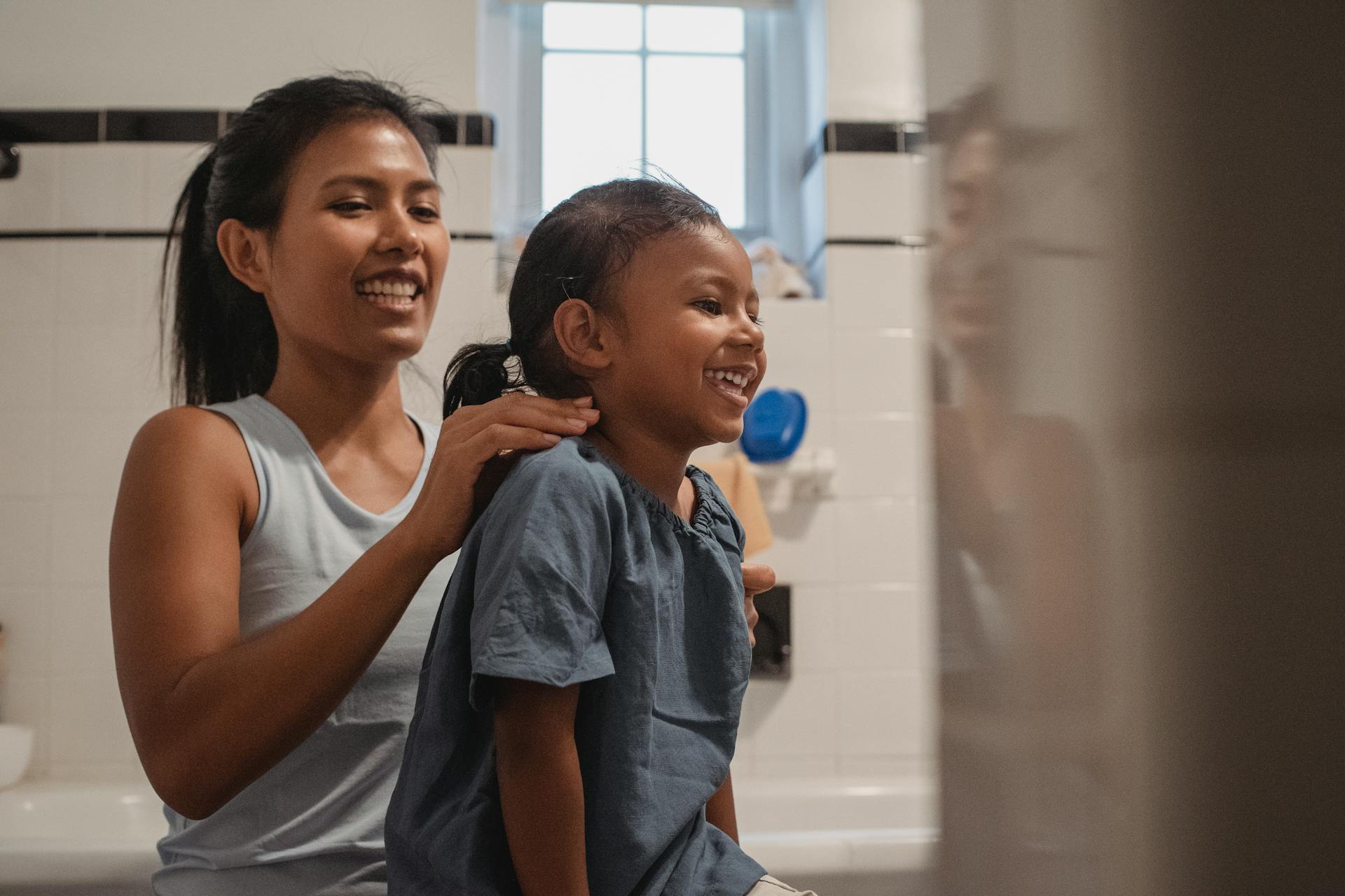 Happy young Hispanic female in casual wear brushing hair of adorable little daughter