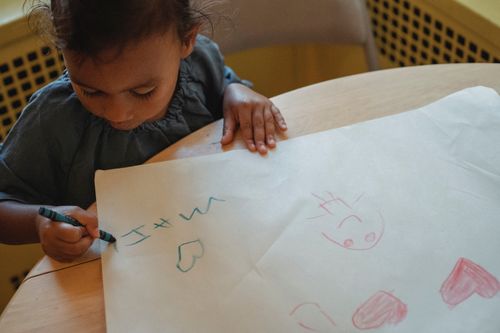 Free From above of concentrated little Hispanic girl drawing with wax crayons on white paper while sitting at table Stock Photo