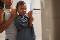Crop anonymous young female in casual wear braiding cheerful little daughter looking at camera