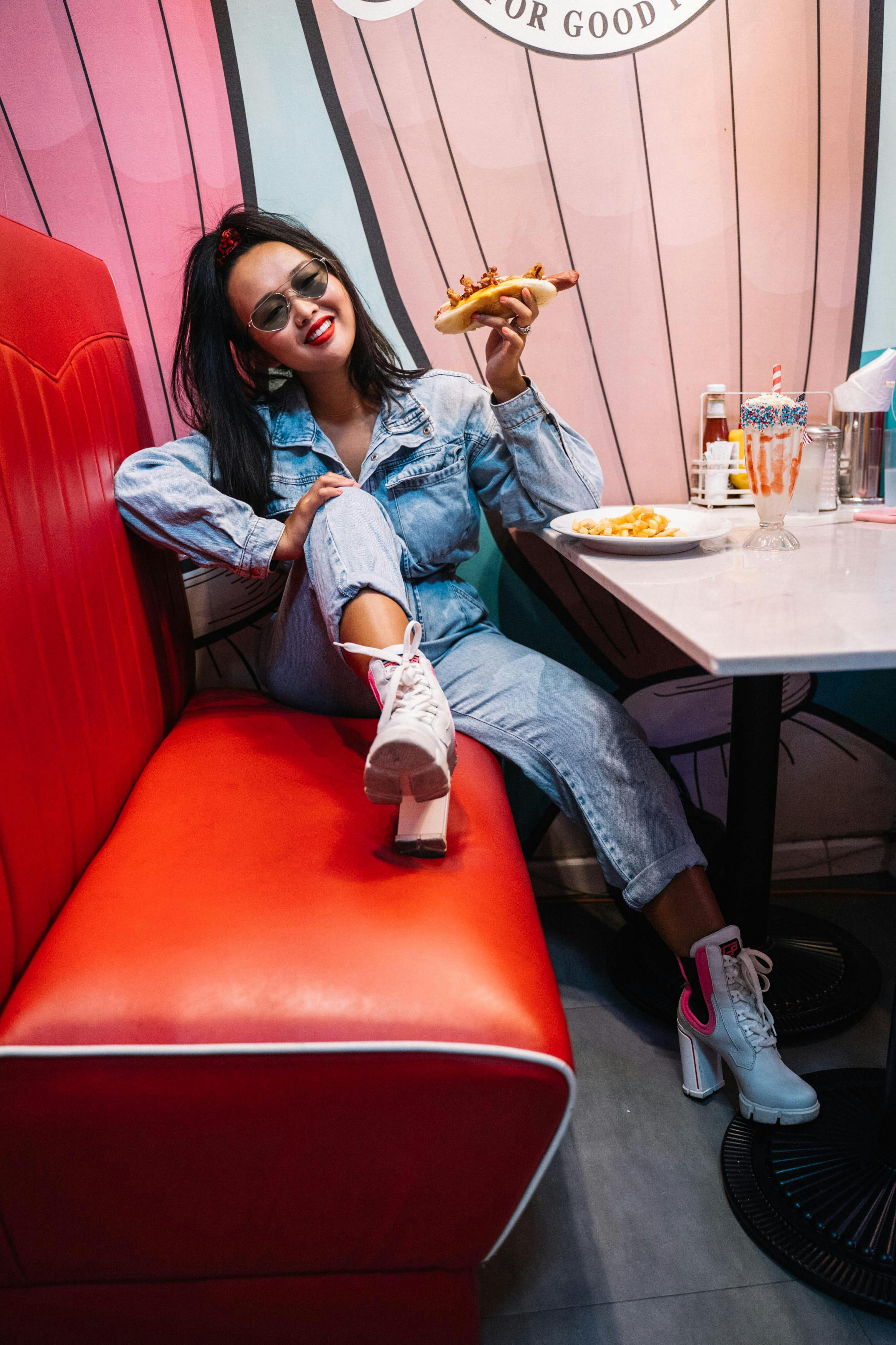 Chic Asian woman in denim sits in a retro diner enjoying a sandwich. Stylish and vibrant atmosphere.