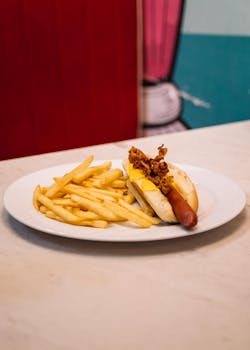 Close-up of a hotdog with cheese and fries on a white plate in a diner setting.