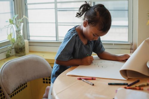 Free Concentrated Hispanic girl in casual clothes sitting at table and drawing picture with wax crayons Stock Photo