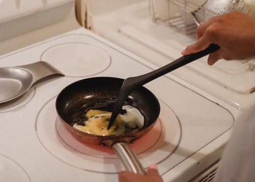 Crop unrecognizable person near white stove with black spatula cooking scrambled eggs in kitchen