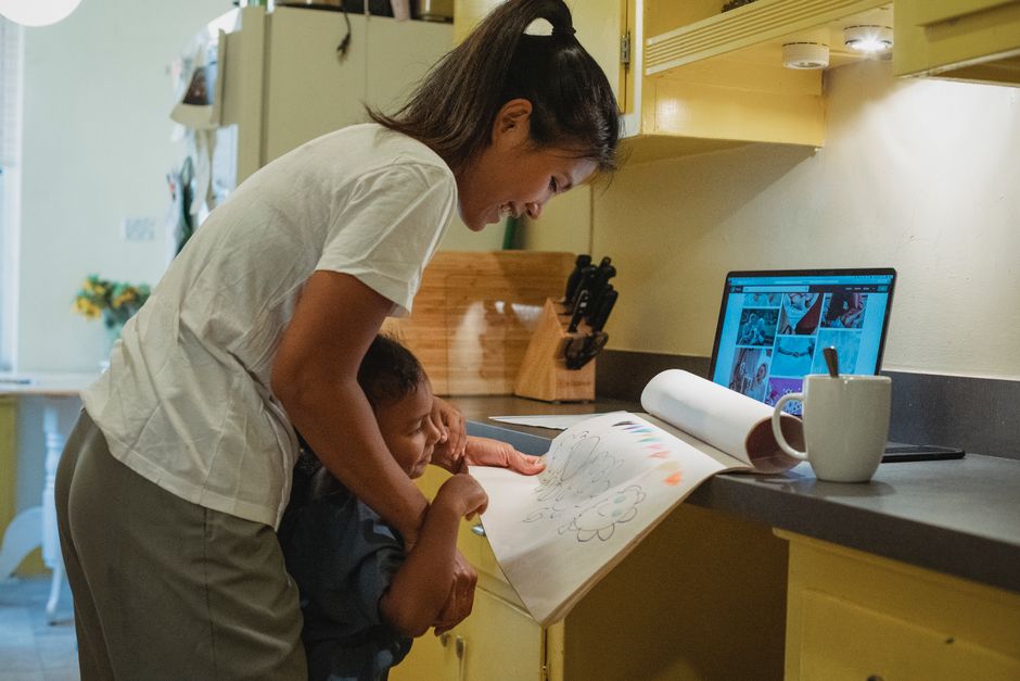 Side view of cheerful young Hispanic female in casual clothes standing with cute daughter showing drawings near table in kitchen
