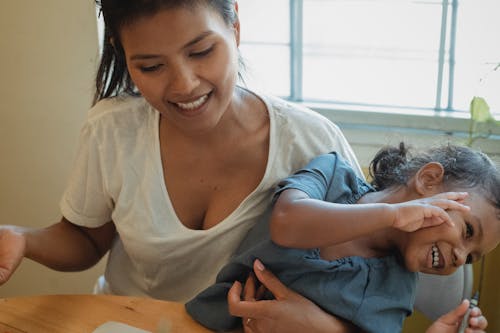 Crop happy ethnic woman smiling and hugging cute joyful little daughter while playing together sitting at table at home