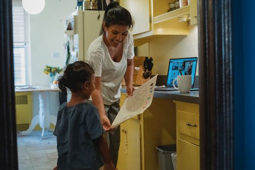 Unrecognizable little ethnic girl demonstrating picture on paper to smiling mother while standing in kitchen with laptop in light apartment