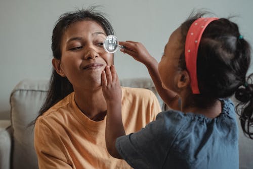 Playful Asian little girl with toy magnifier glass making check up of eye of mother while playing doctor in apartment