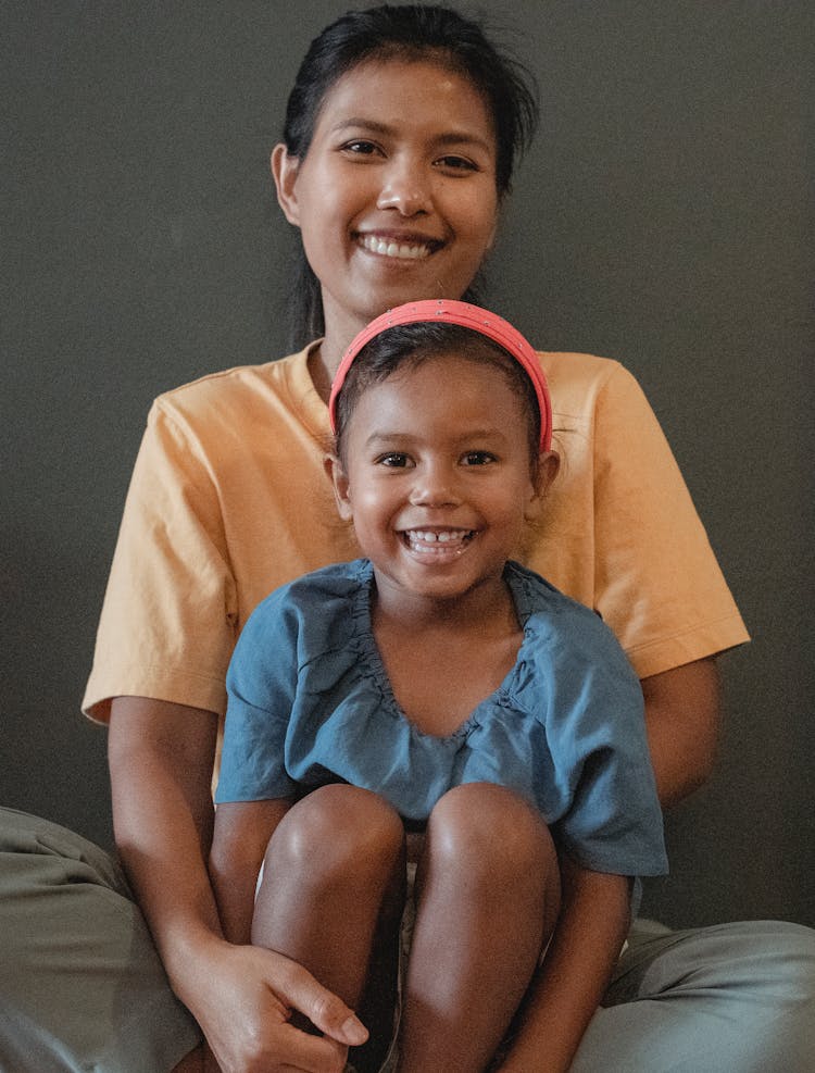 Smiling Asian Mother And Daughter Sitting On Couch At Wall