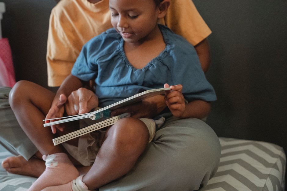 Unrecognizable ethnic mother hugging little girl and reading interesting book while sitting on couch near wall during weekend in apartment