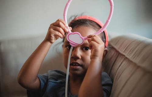 Pretty ethnic little girl looking at camera while sitting on couch with raised arms and pink toy stethoscope in hands