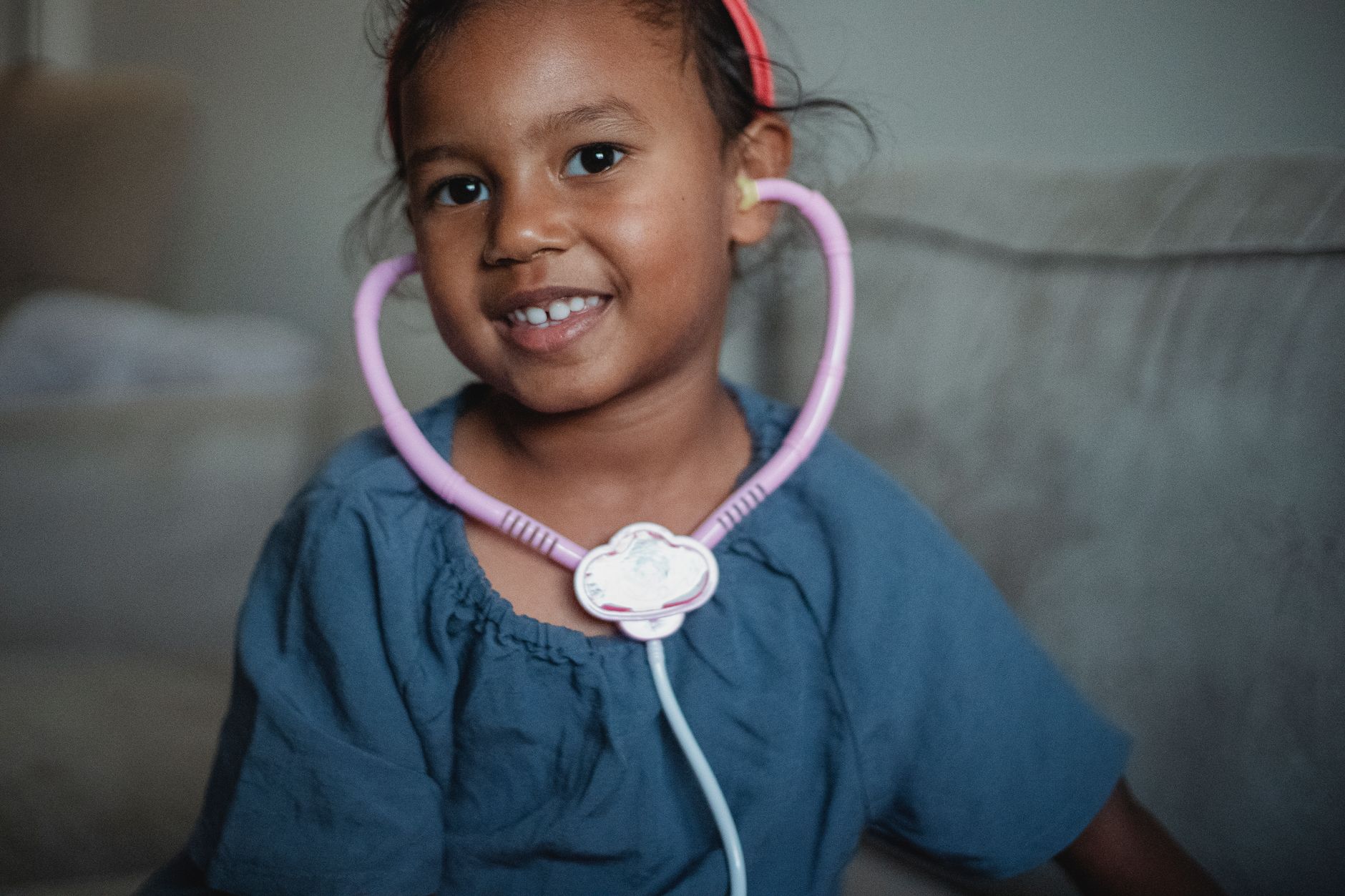 Positive ethnic little girl with toy stethoscope looking away while sitting on couch and playing doctor on blurred background at home
