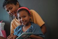 Cheerful ethnic mother hugging positive little girl looking at camera while reading book near black wall during weekend in apartment