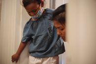 Caring Asian mother helping crop little girl with protective mask while standing in doorway in apartment before going on street