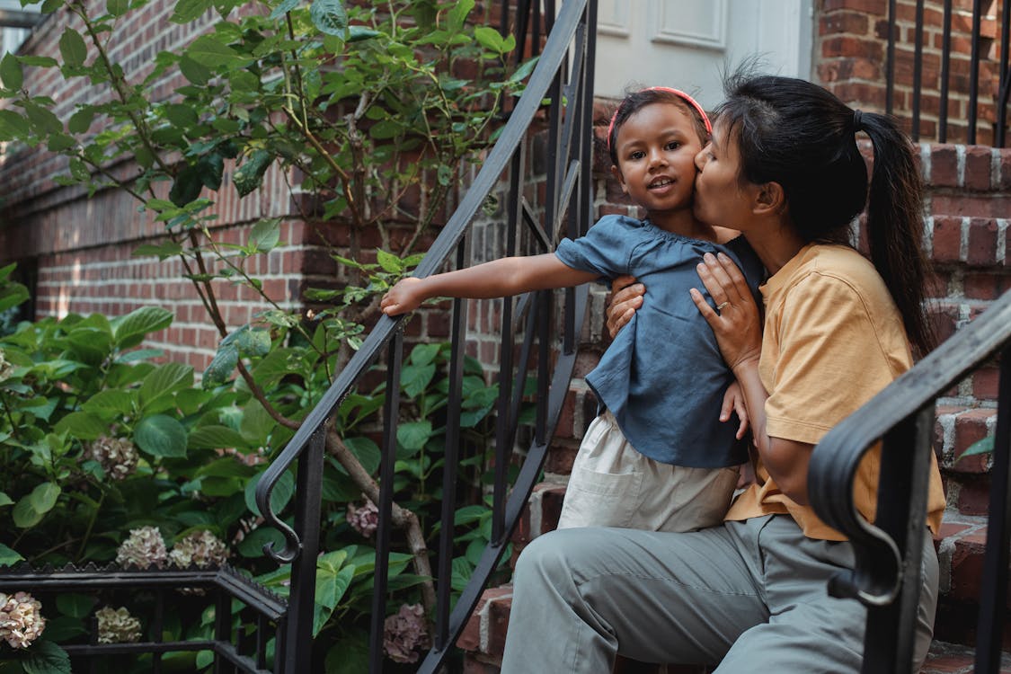 Mother hugging and kissing little girl while sitting on stairs of house with metal railing