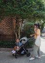 Full body of focused Asian mother with bag talking on smartphone while walking on pathway with happy little girl sitting in baby stroller