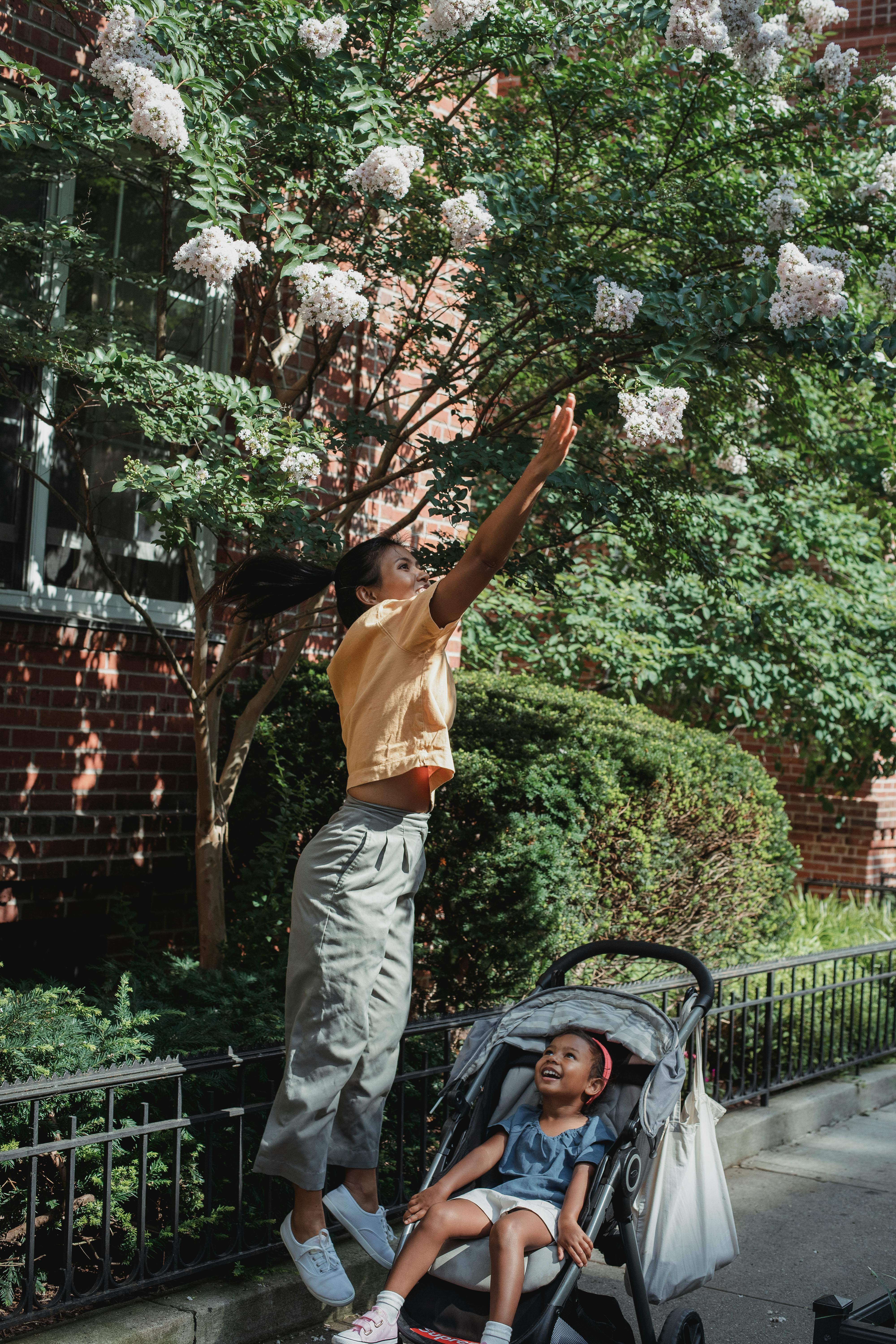 ethnic woman reaching for flower on street with daughter in carriage