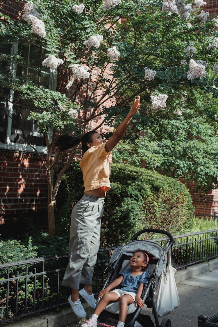 Ethnic Woman Reaching For Flower On Street With Daughter In Carriage