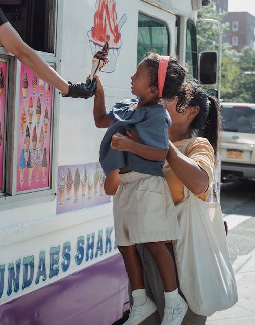 Faceless mother holding little girl while standing near food truck and buying chocolate ice cream on city street