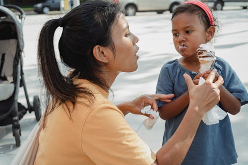 Attentive ethnic mother helping cute funny little daughter with tasty sweet ice cream cone on street in summer