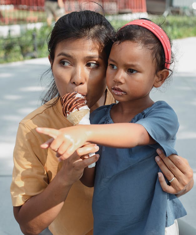 Young ethnic mother with adorable small daughter tasting sweet delicious ice cream cone and looking away together on street