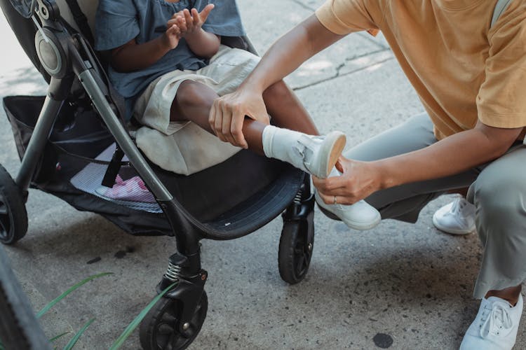 Ethnic Mother Helping Daughter Putting On Shoes In Baby Carriage