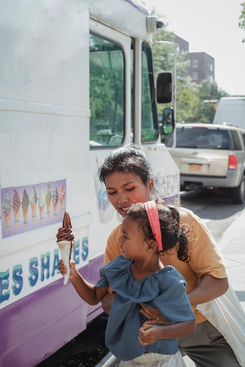 Tender ethnic mother hugging cute little daughter with delicious ice cream cone near wagon with desserts