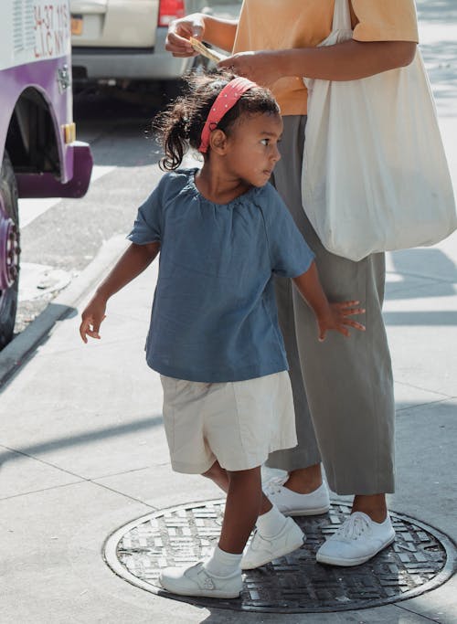 Free Adorable small ethnic girl in summer outfit near anonymous mother on street in sunny weather Stock Photo