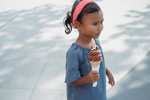 Crop little adorable ethnic girl in casual outfit with tasty sweet ice cream on street in summer