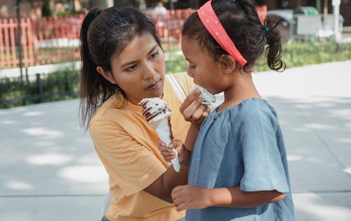 Ethnic caring mother wiping face of adorable crop daughter with sweet tasty ice cream cone
