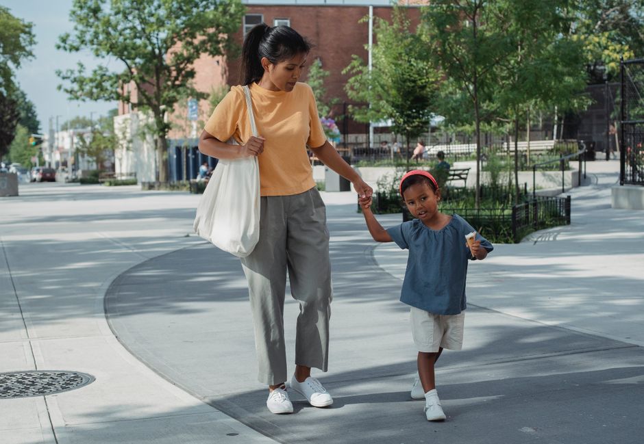 Full body of calm Asian mother in casual clothes holding hand of little daughter while walking on street in city in sunny day