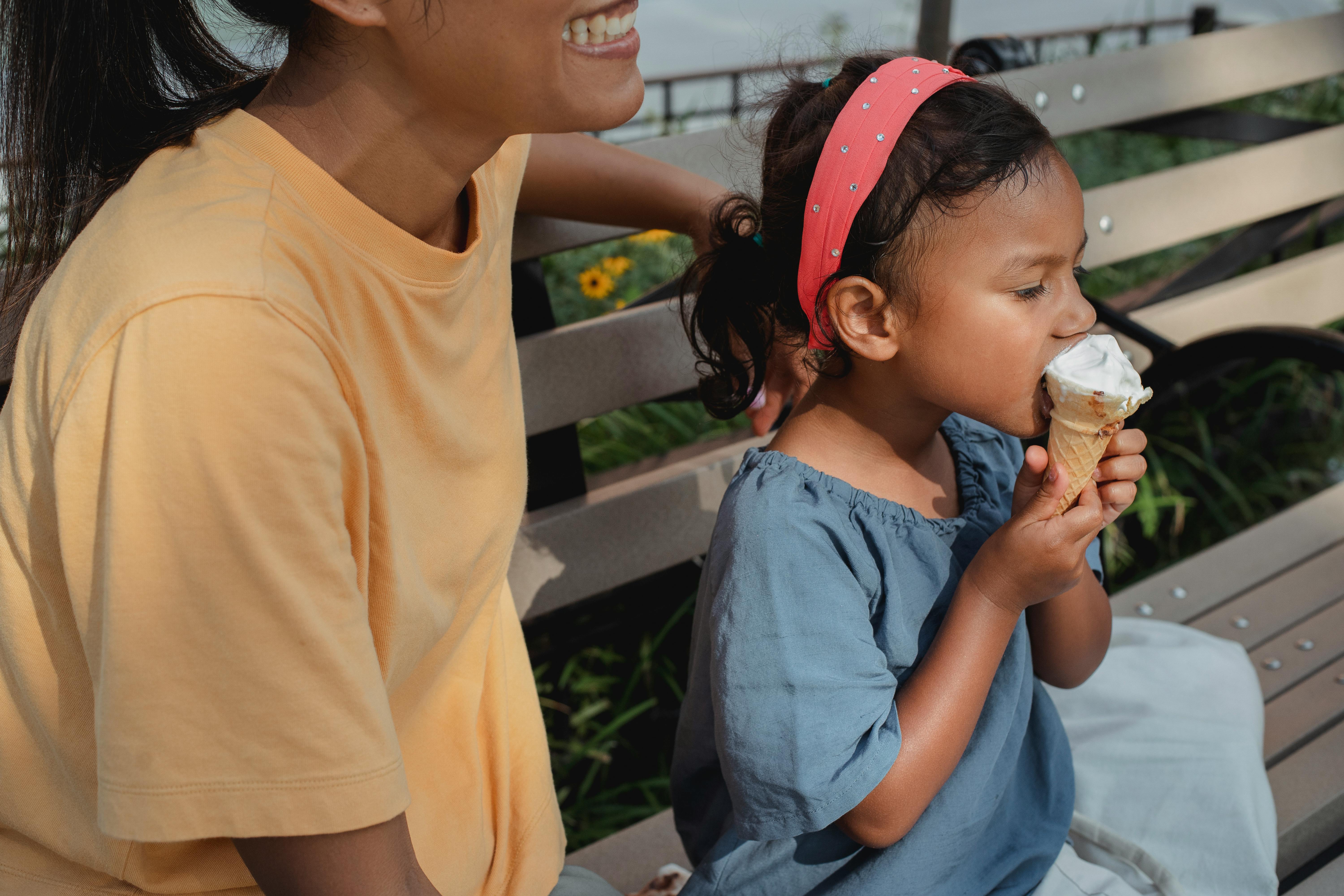 positive woman sitting on bench near little girl eating ice cream