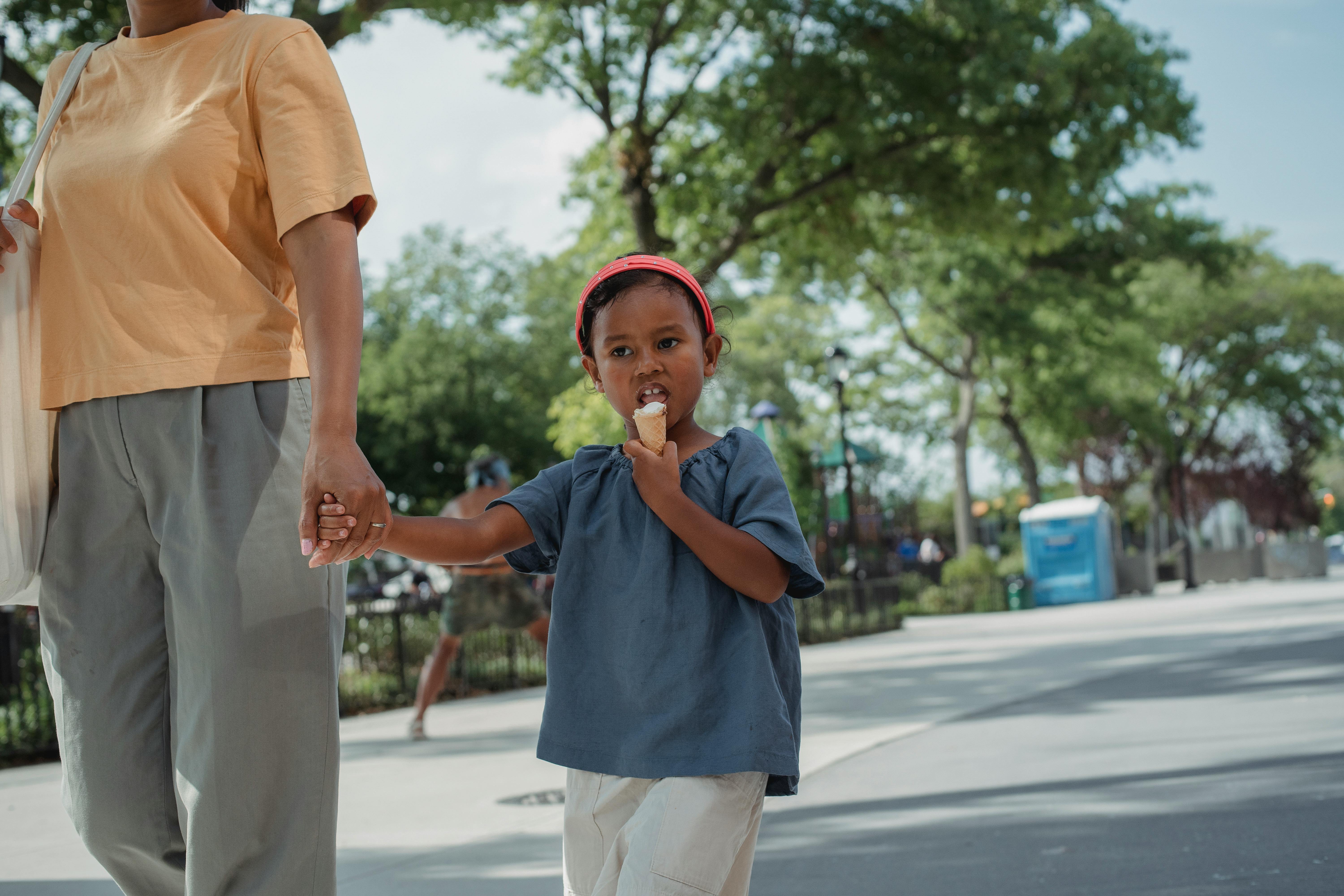 ethnic child eating tasty ice cream and walking on street with mother