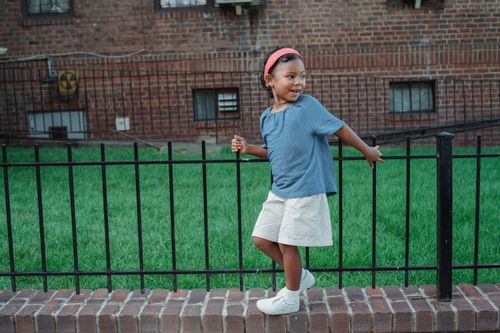 Free Full length of positive Asian child with dark hair in casual clothes standing with raised hands near metal fence against building and looking away in daytime Stock Photo