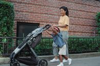 Side view of cheerful Asian woman in casual clothes walking small daughter rolling baby carriage on street near brick building in daytime