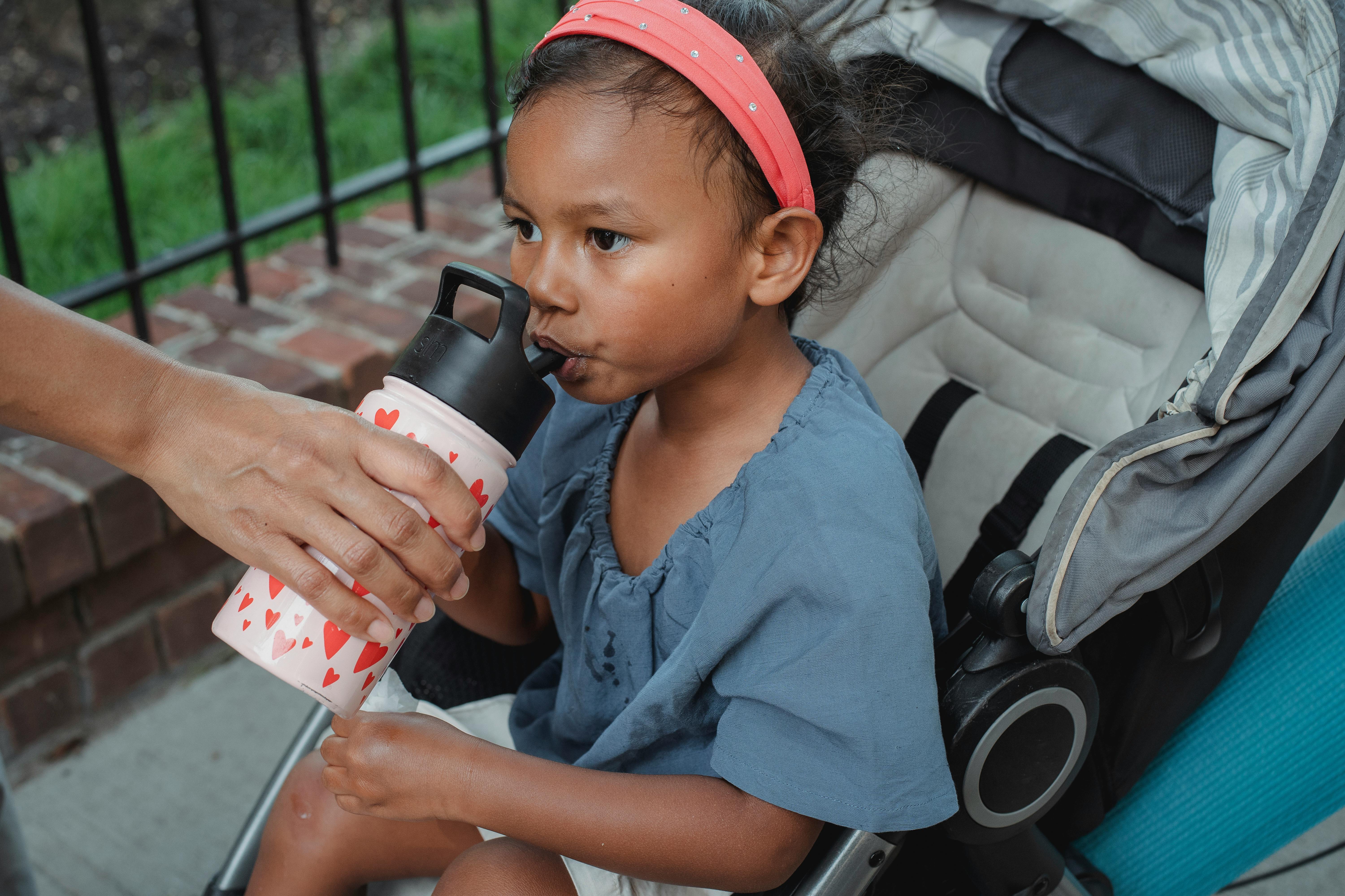 small ethnic girl sitting in stroller and drinking beverage from bottle