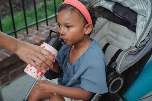 Calm Asian girl with brown eyes and dark hair in casual clothes sitting in stroller and drinking water from bottle from mother hand while spending time on street in daylight