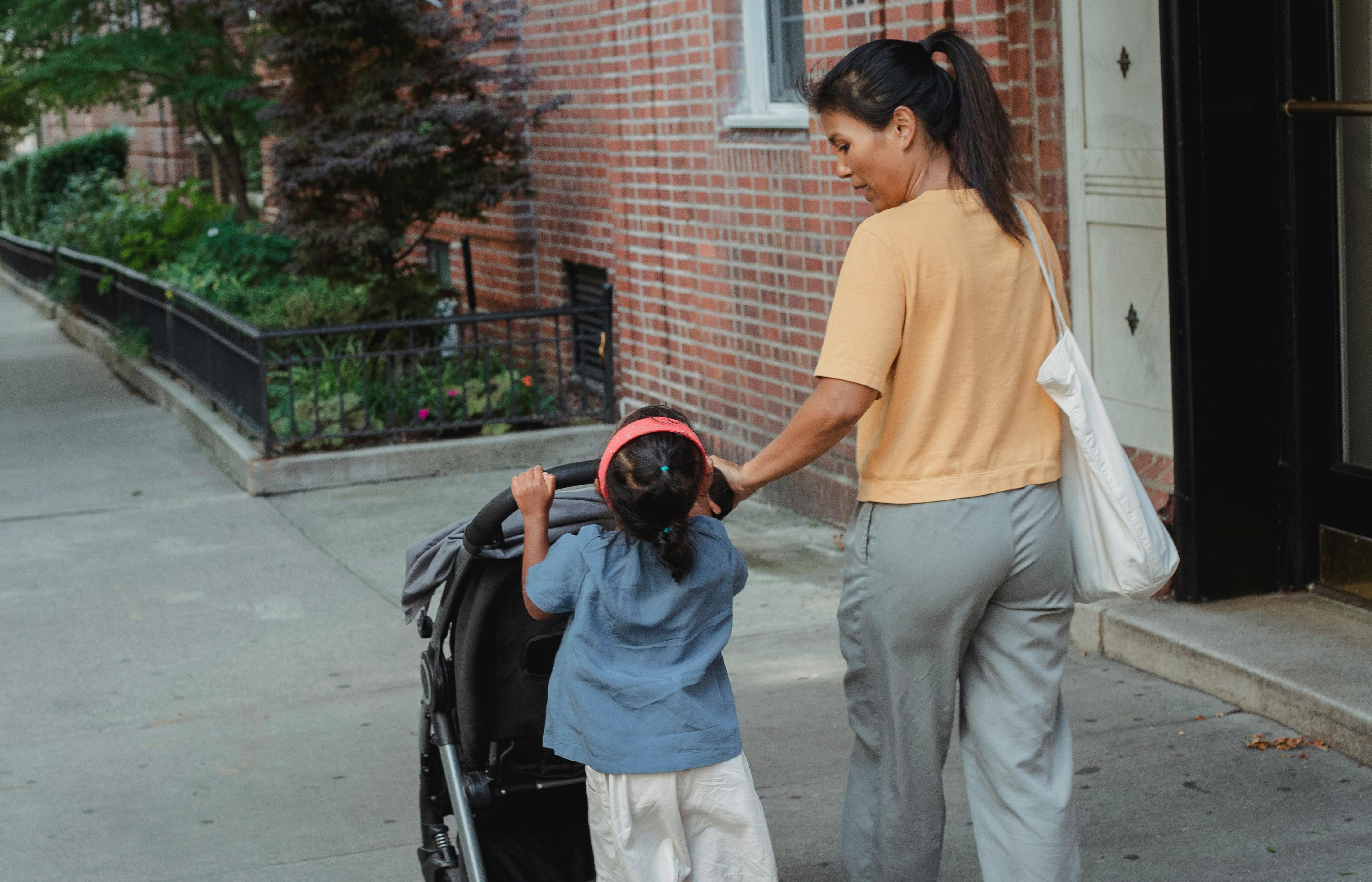 Back view of ethnic woman carrying stroller with little girl on sidewalk of street