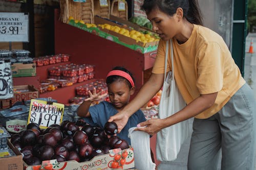 Free Asian woman putting black plum into eco bag while choosing fruits from box in street market with daughter Stock Photo