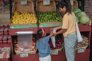 Back view of Asian girl picking ripe limes from box to eco friendly bag while shopping with mother in market